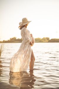 Werdende Mutter steht mit einem Spitzenkleid zum Sonnenuntergang im Wasser und haelt ihren Babybauch.